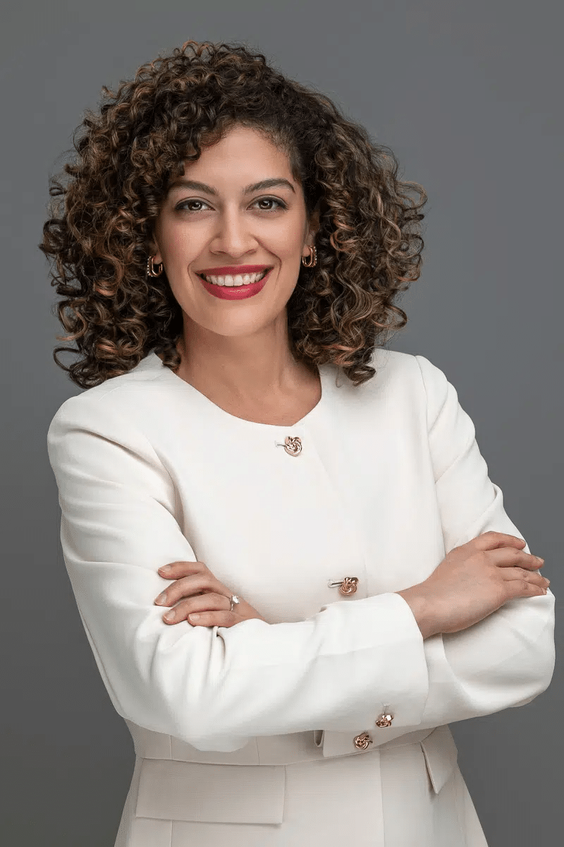 Professional studio headshot of partner attorney B. Nina Vazquez with curly hair, smiling and wearing an elegant off-white structured blazer with gold buttons against a solid gray backdrop.