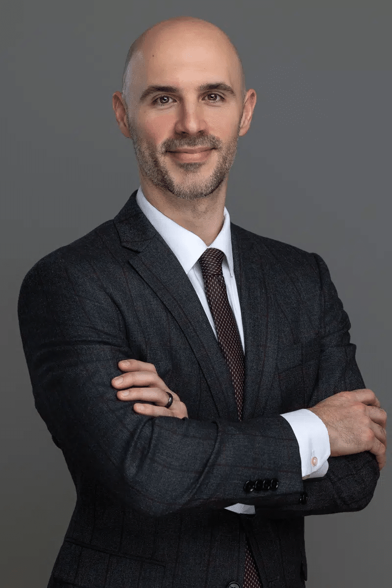 Professional studio headshot of partner attorney Stefano Mittone wearing a dark plaid suit, white dress shirt, and a textured dark red tie with his arms crossed against a gray background.